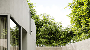 Modern concrete home exterior with large windows and lush green trees in backyard garden setting