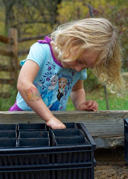 Child planting in a black plastic seedling tray outdoors near a wooden garden bed