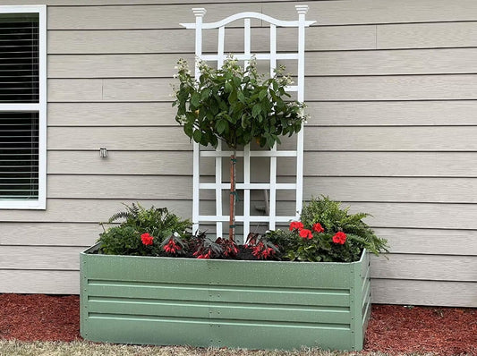 Green metal raised garden bed with trellis, red flowers, and green plants against beige siding