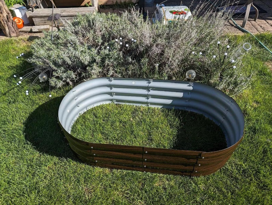 Oval metal raised garden bed with wood base, set in green lawn beside shrubs and garden decor
